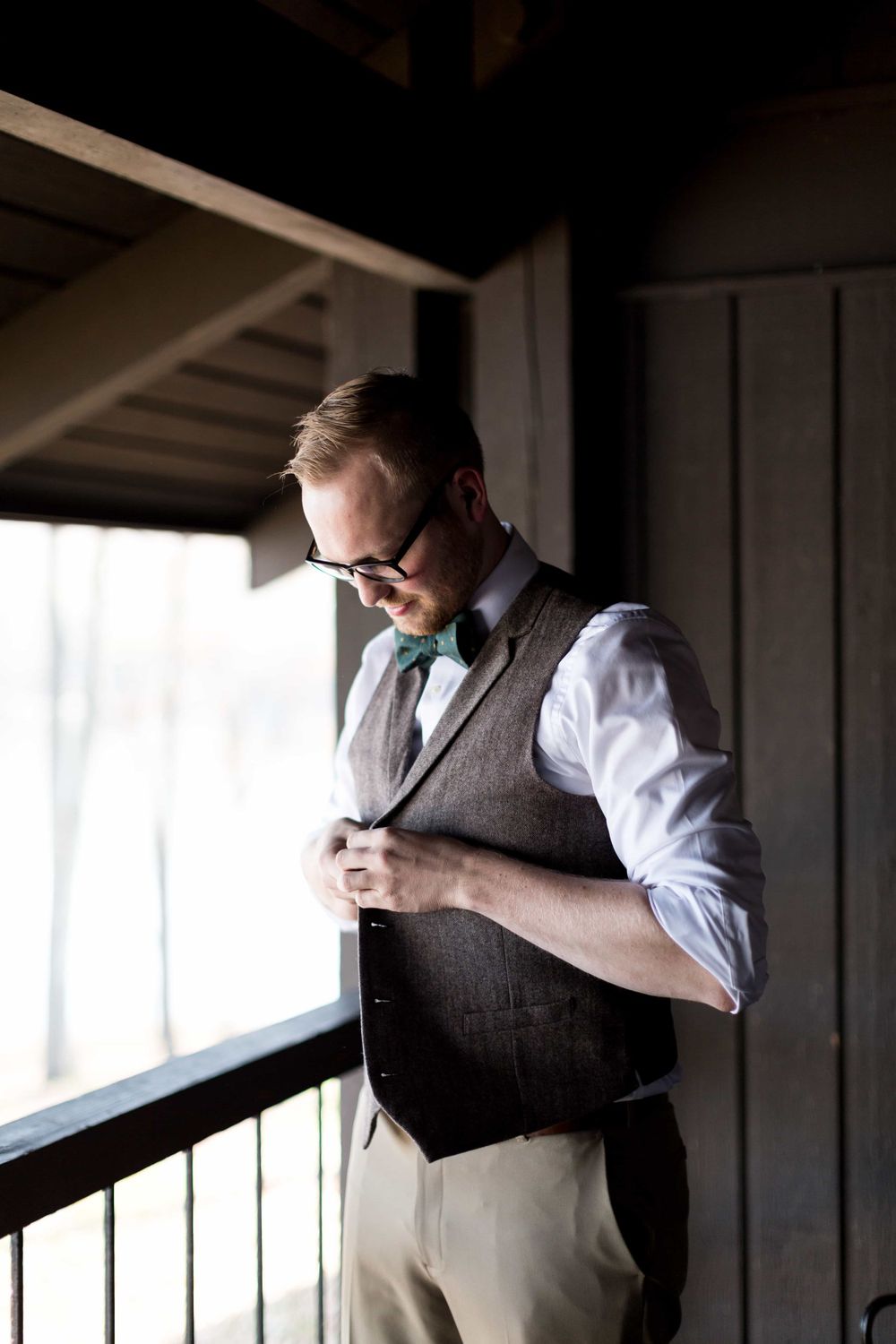 Dapper groom in gray tweed vest and teal bow tie buttoning up near a window.