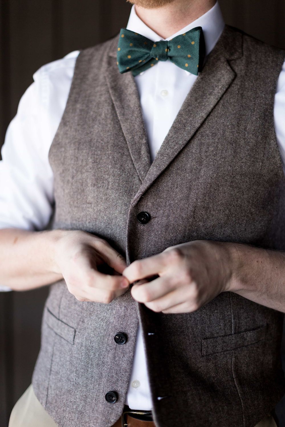 Close-up of groom in gray tweed vest fastening buttons with teal polka dot bow tie.