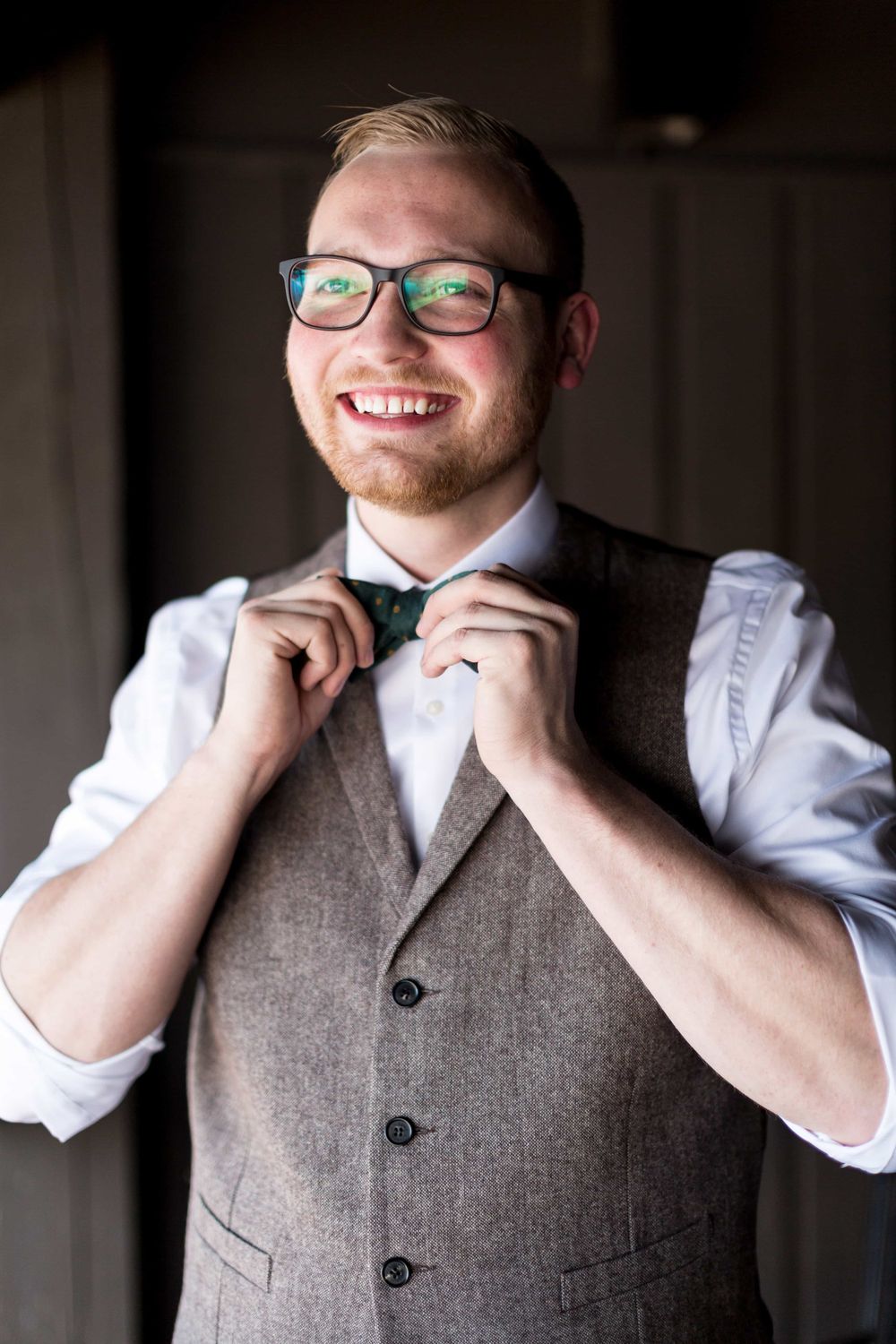 Smiling groom with glasses and red beard adjusting his green polka dot bow tie.
