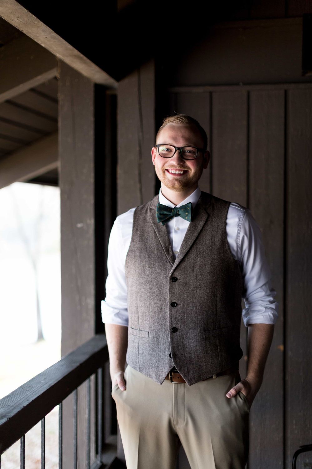 Smiling groom in brown tweed vest, green bow tie, and glasses standing on a rustic wooden porch.