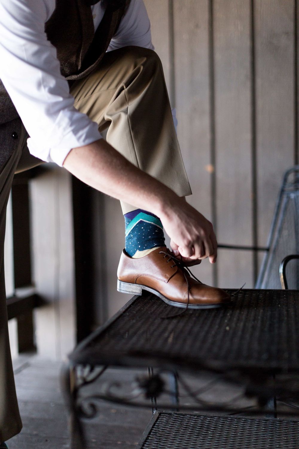 Groom tying brown leather dress shoes with colorful teal patterned socks on a dark bench.
