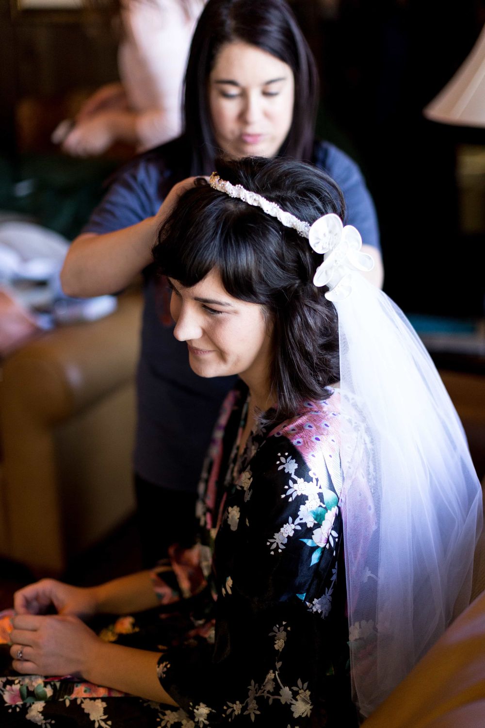 Bride with dark hair having floral wedding headpiece and veil adjusted by bridesmaid before ceremony.