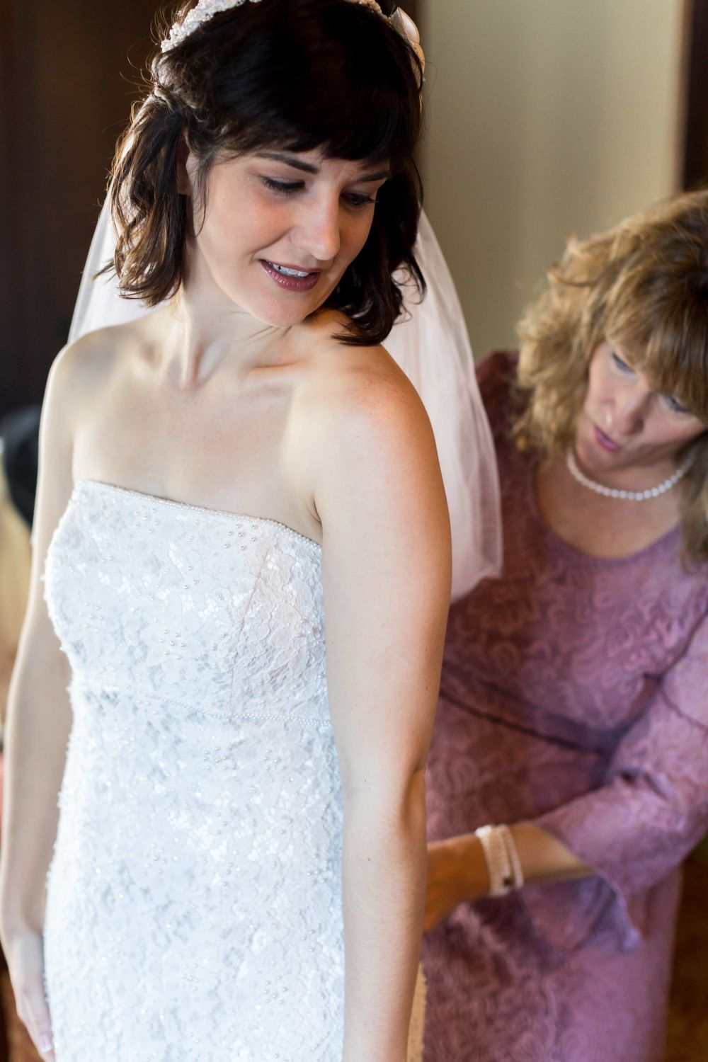 Bride in strapless lace wedding gown being helped by older woman in pink dress.