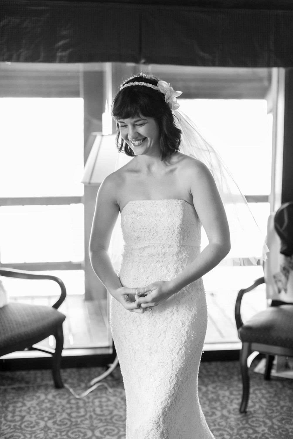 Black and white photo of radiant bride in strapless lace gown smiling near bright windows.