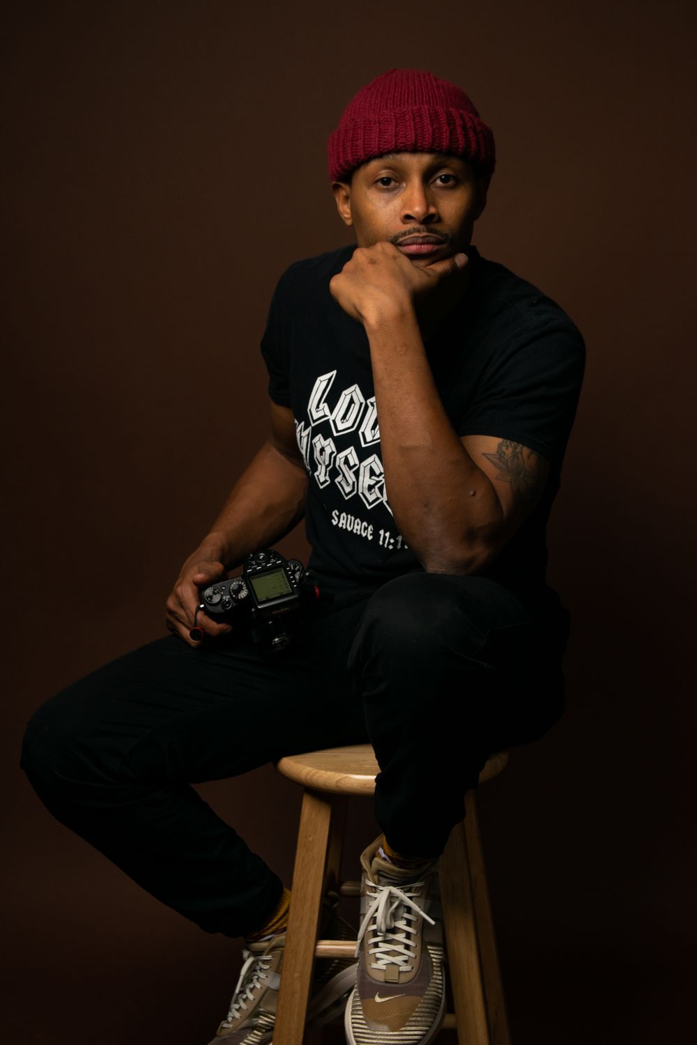Portrait in red beanie and black t-shirt sitting on wooden stool against brown backdrop.