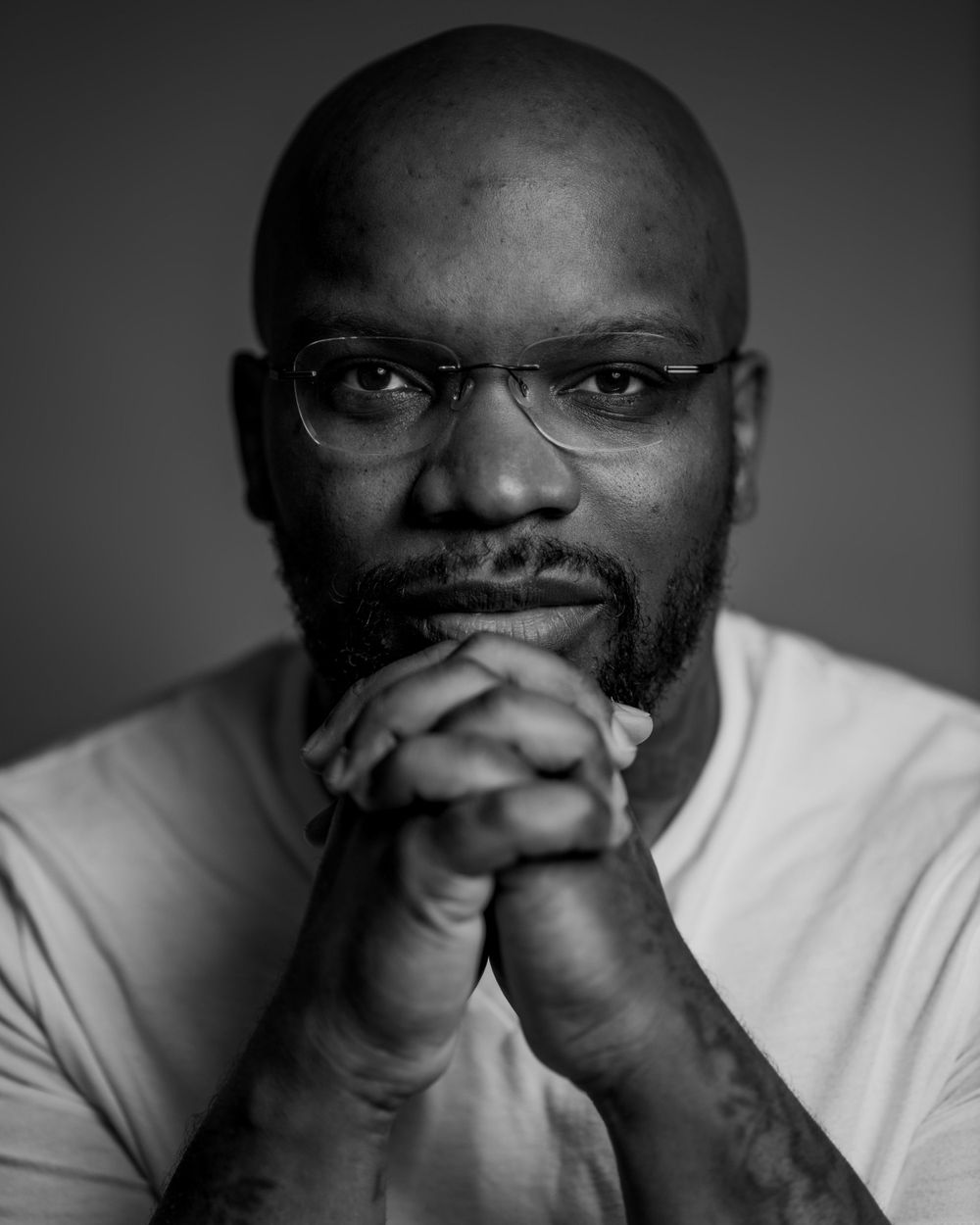 Black and white portrait with clasped hands and glasses against dark background.