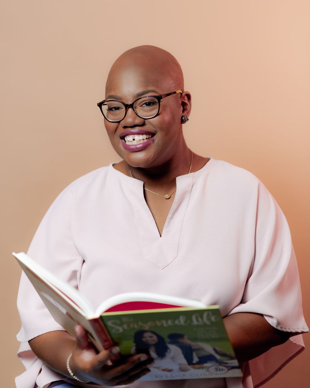 Cheerful reader in glasses and pink top holds open book while smiling at camera.