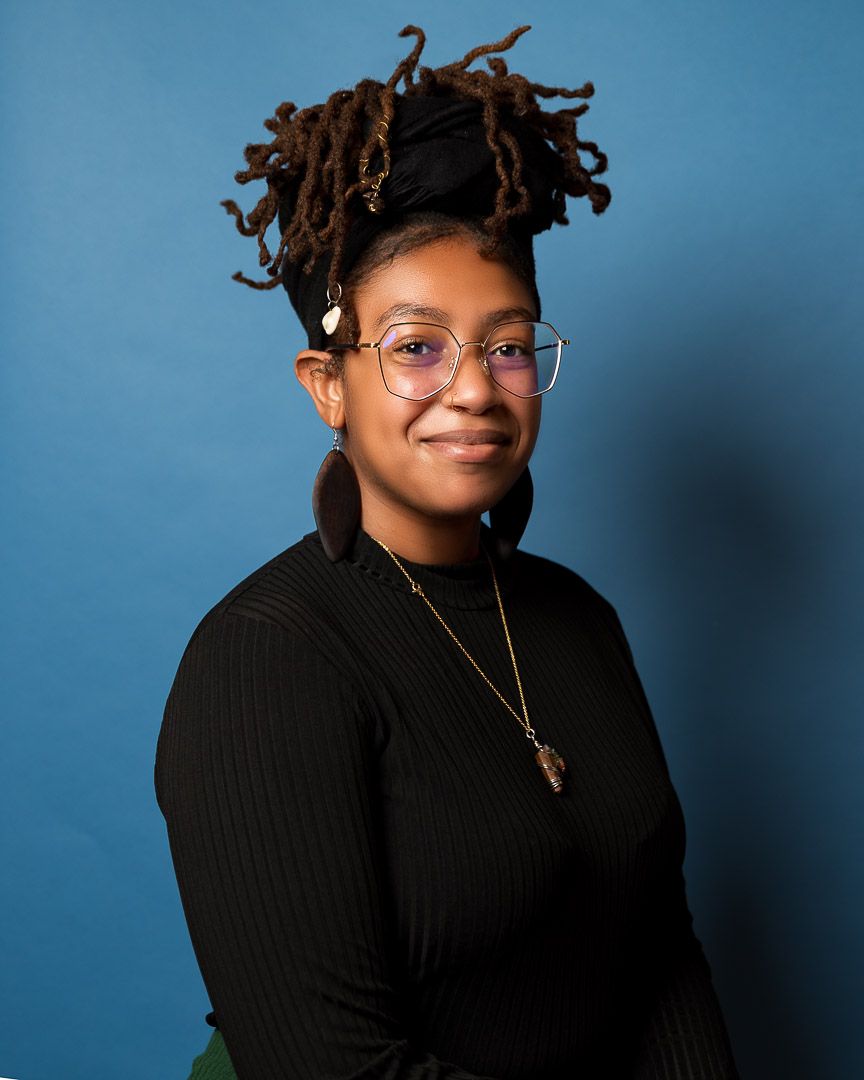 Portrait of someone in glasses with styled locs against a blue backdrop wearing a black sweater.