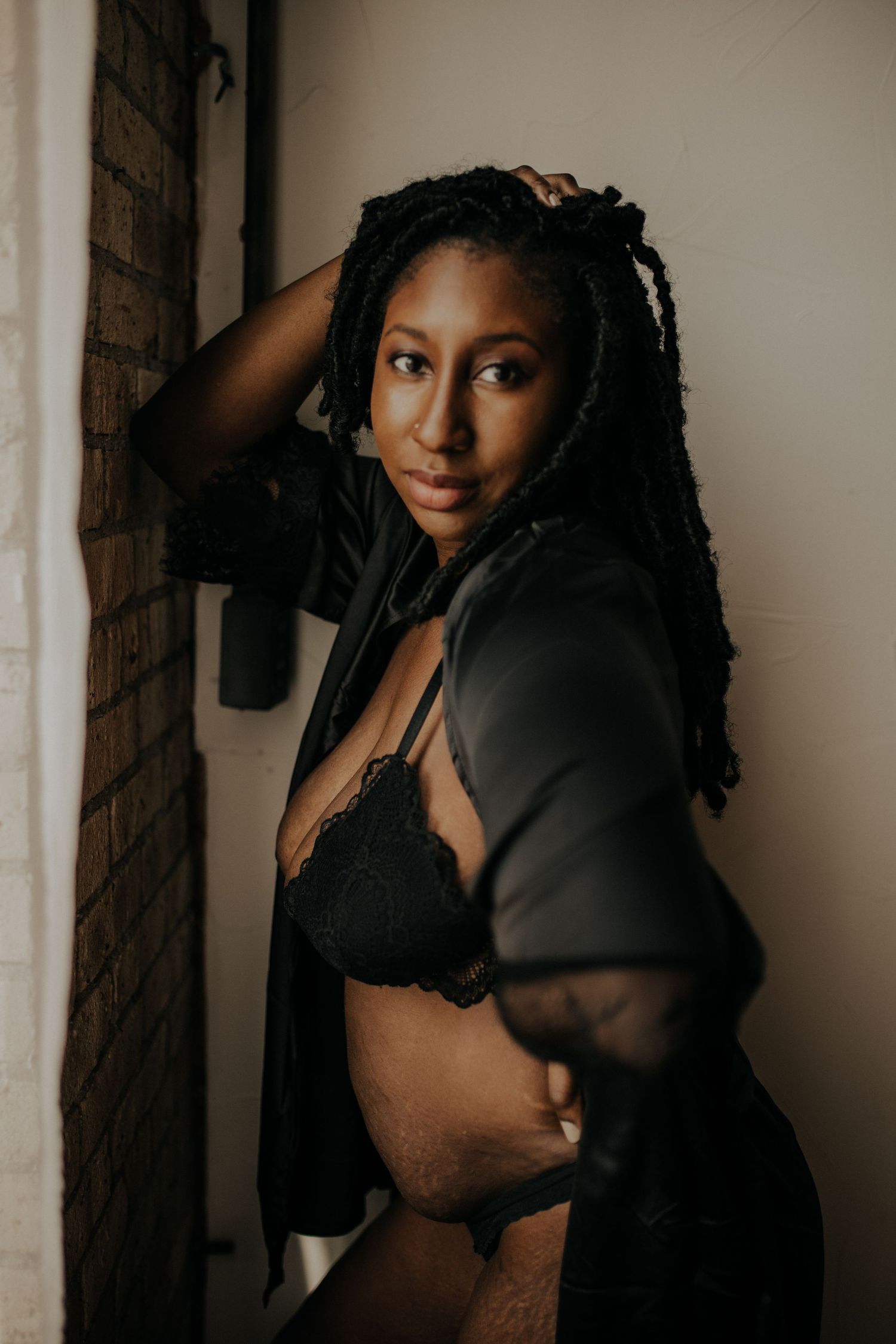 Artistic boudoir portrait of woman in black lingerie and sheer robe against brick wall in warm moody lighting.