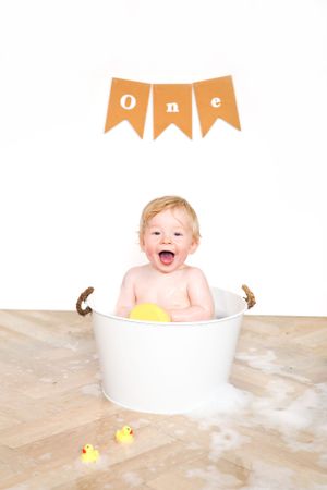 Smiling baby sits in white bucket with rubber ducks under orange 'one' banner.