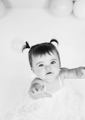 Black and white portrait of baby with pigtails in white dress against light background.