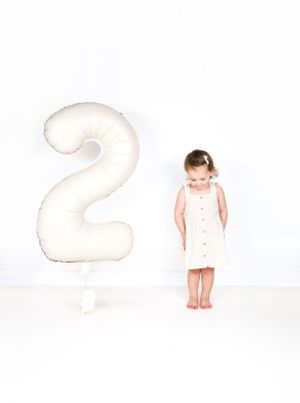 Small child in white dress stands next to large white number 2 balloon against plain background.