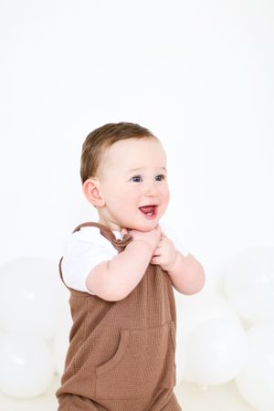 A happy toddler in brown overalls poses against a white background with balloons.