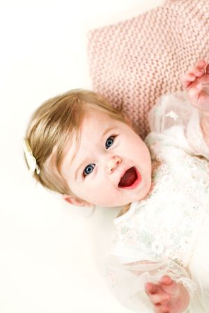 A joyful baby in white lace lying on a pink knit blanket.
