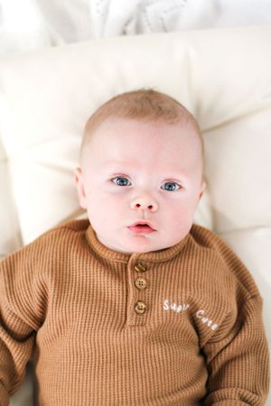 A baby in a brown knit sweater lies on white bedding.