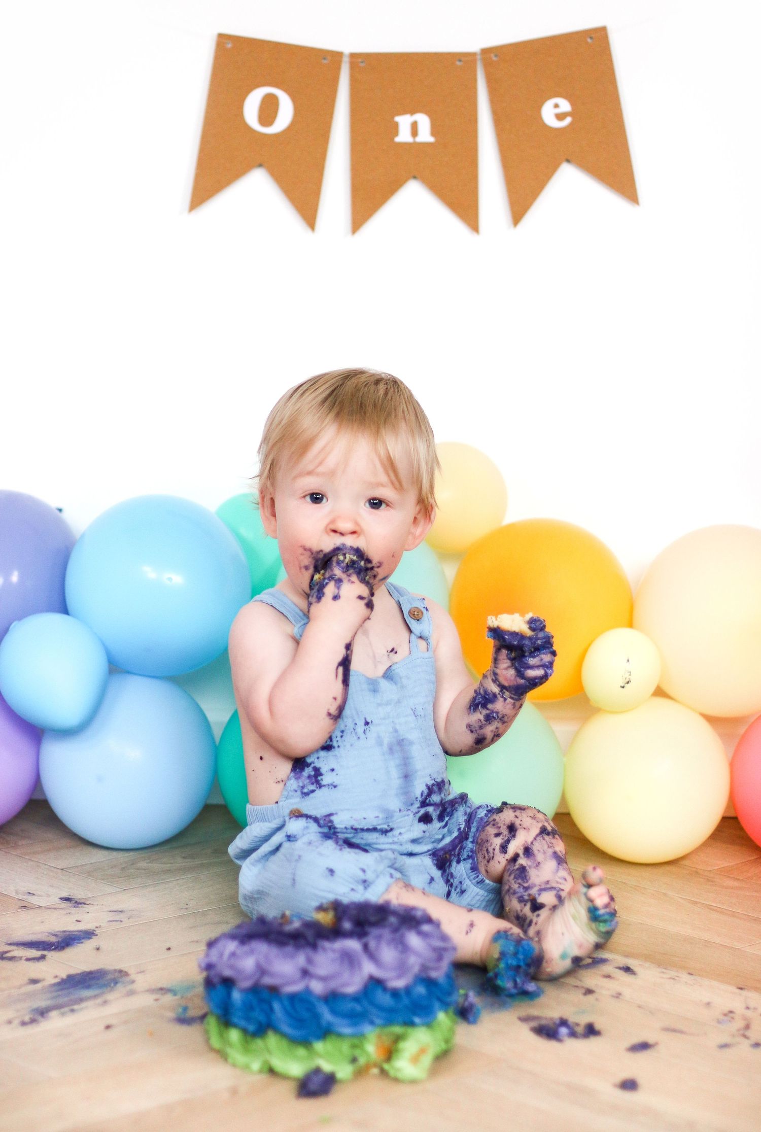 A messy baby covered in purple cake sits among rainbow balloons under a 'One' banner.