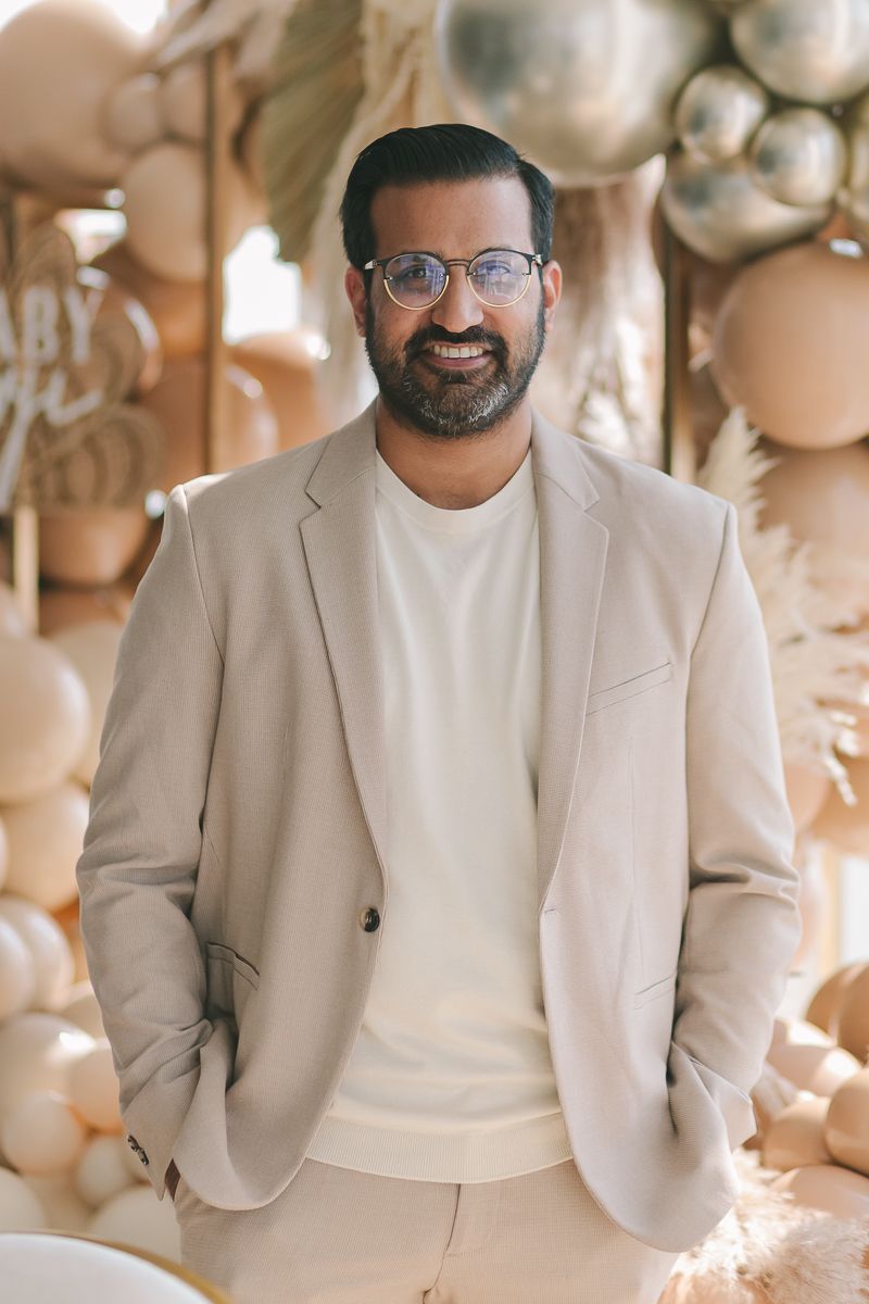 a half body view of an Indian man wearing a light suit and cashmere sweater with balloons in the background by Paula O