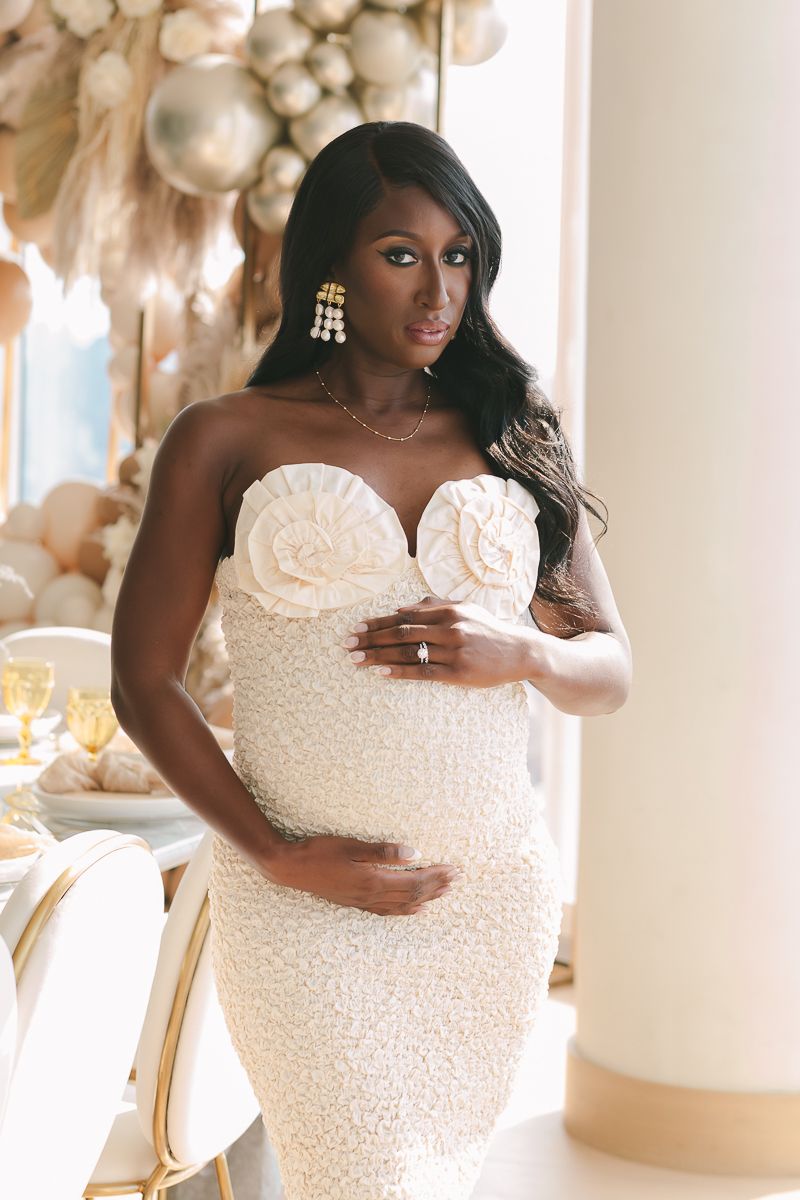a half body view of a black pregnant woman wearing a white dress with balloons in the background during her baby shower