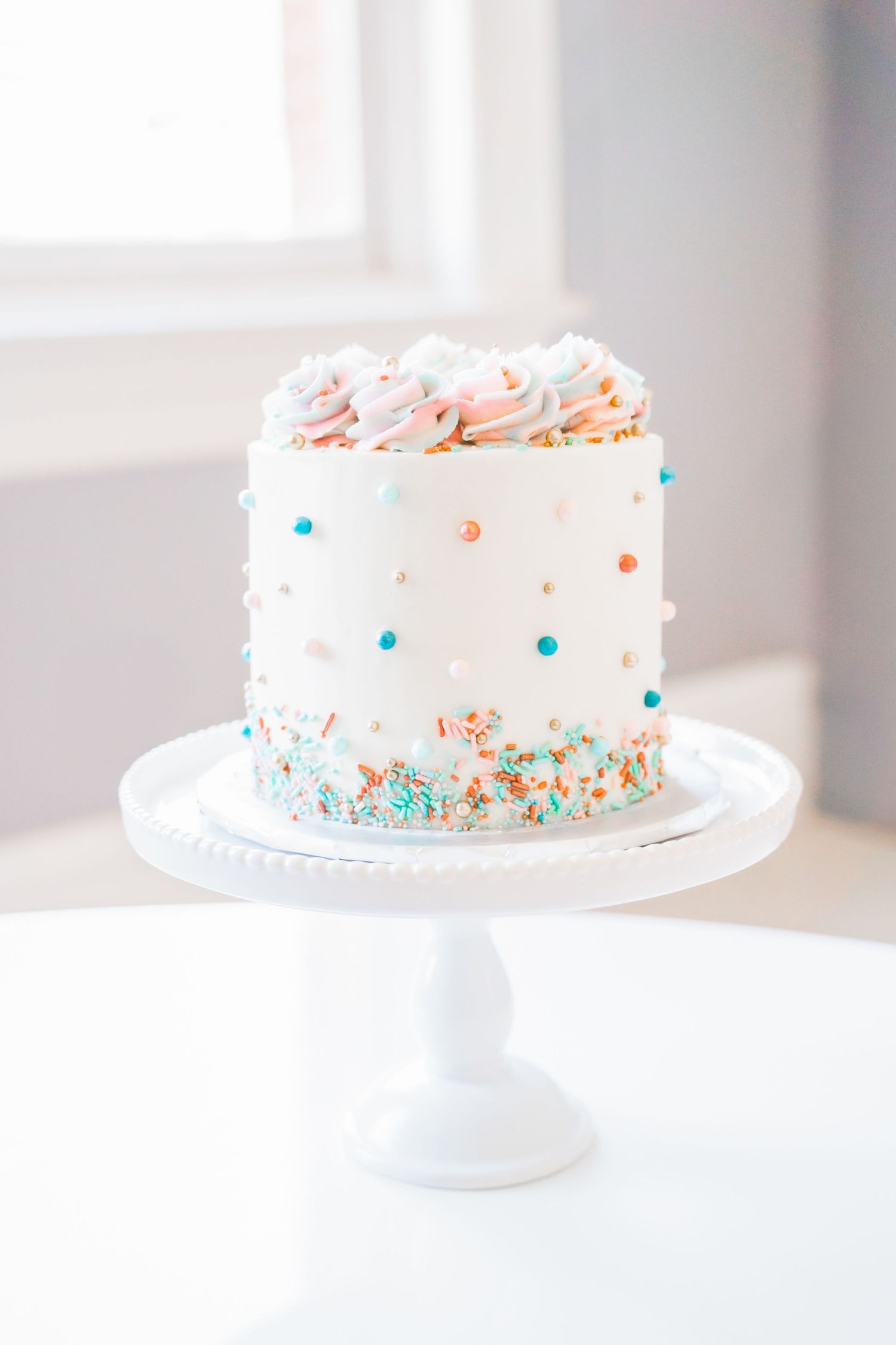 White layered cake with colorful sprinkles and polka dot decorations displayed on a white pedestal cake stand.