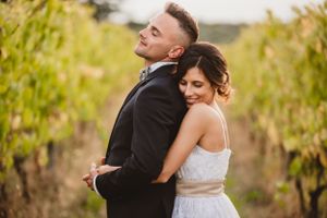 A romantic couple poses together in a vineyard at sunset wearing wedding attire.