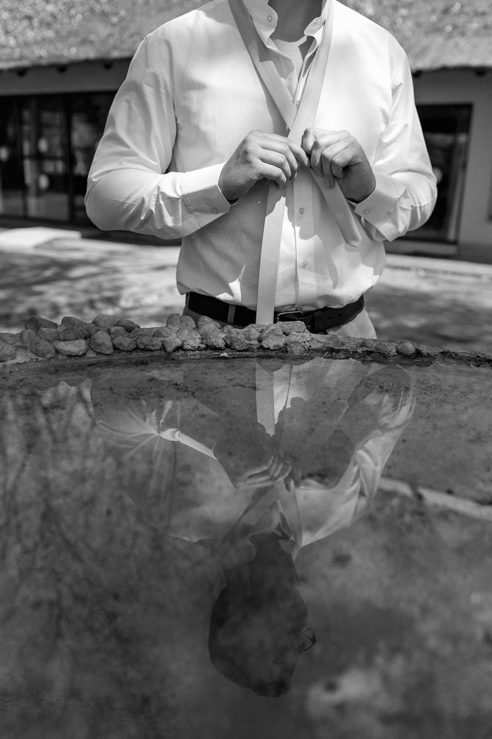 Groom tying his tie while his reflection shows in front of him.