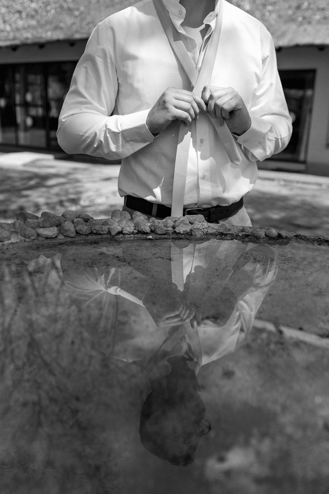 Groom tying his tie while his reflection shows in front of him.