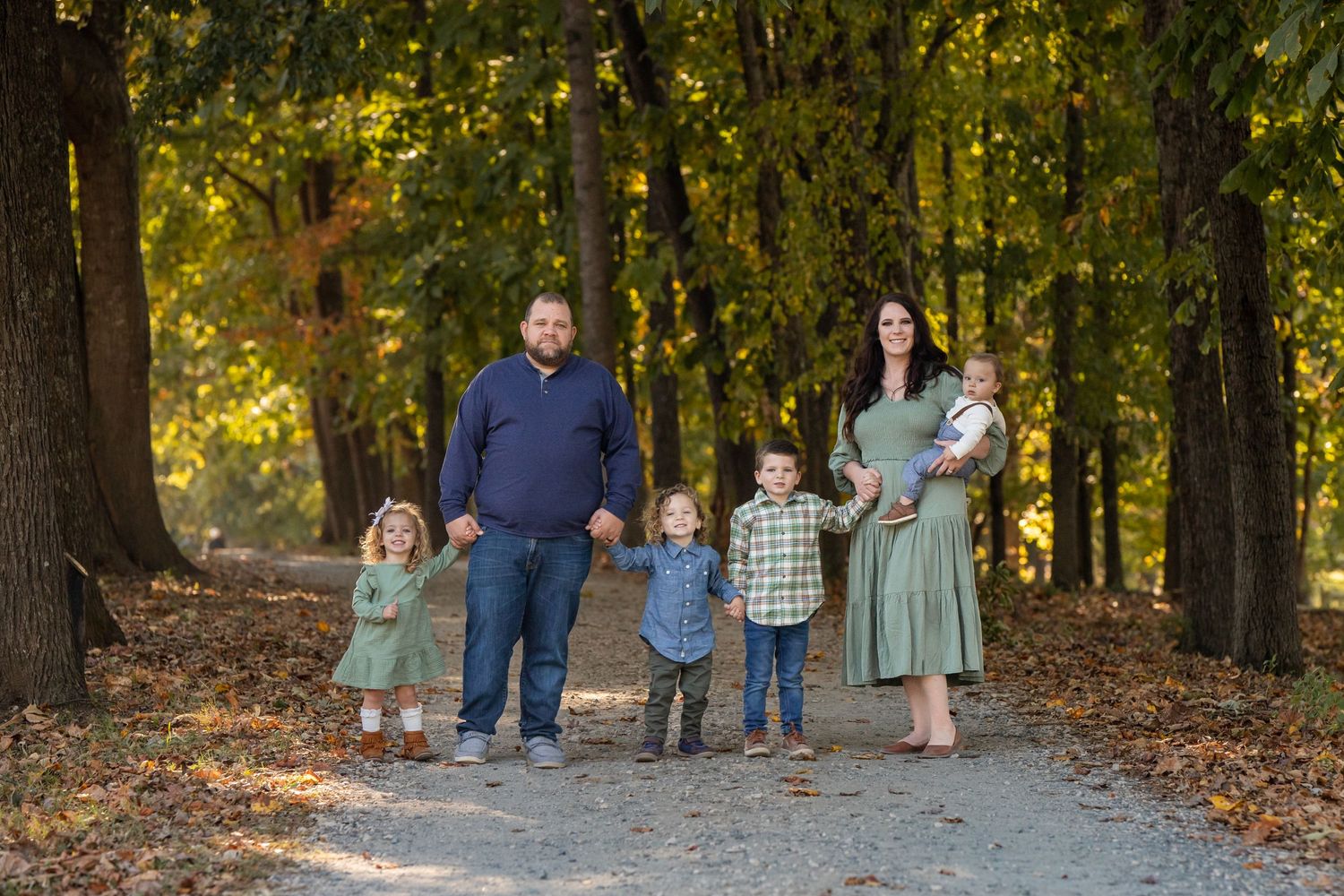 A family stands together on a tree-lined path covered in fallen autumn leaves during an outdoor photo session.