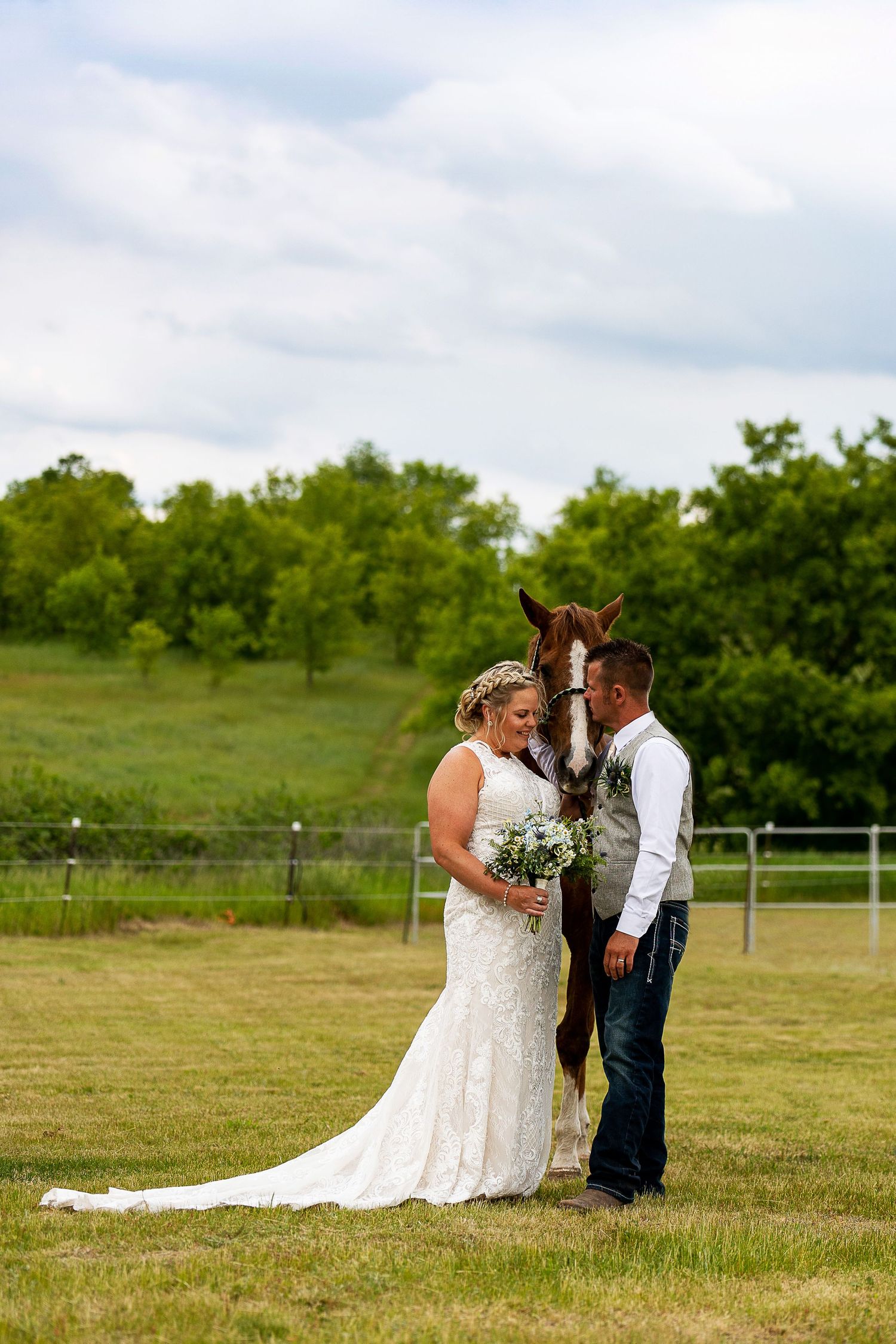 Bride and groom share a romantic moment with a brown horse on a green farm during their outdoor wedding.