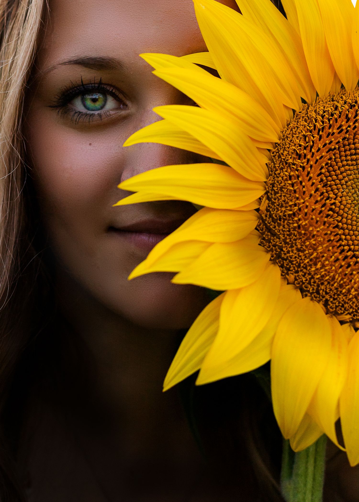 Close-up portrait of young woman with green eyes partially hidden behind vibrant yellow sunflower.