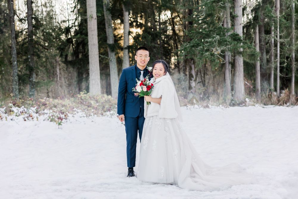 Bride and groom posing together in snowy woodland setting, bride holding red bouquet in elegant white cape.