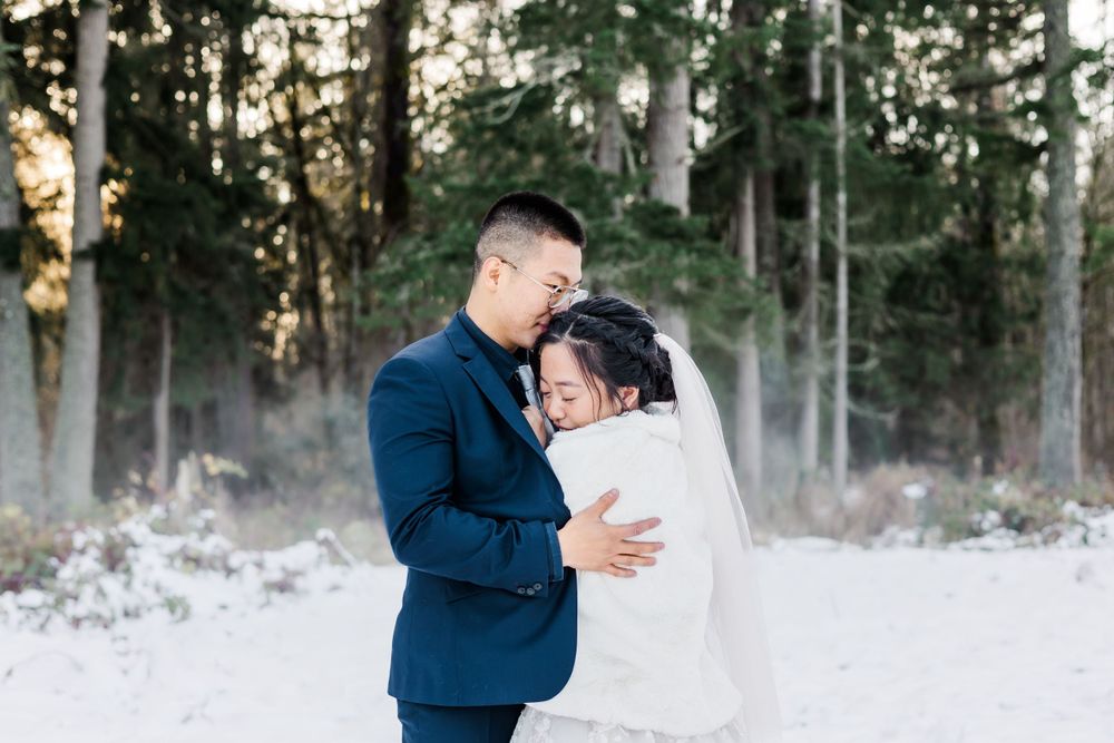 Newlyweds sharing tender forehead touch embrace in snow-covered forest, groom in navy suit and bride in white.
