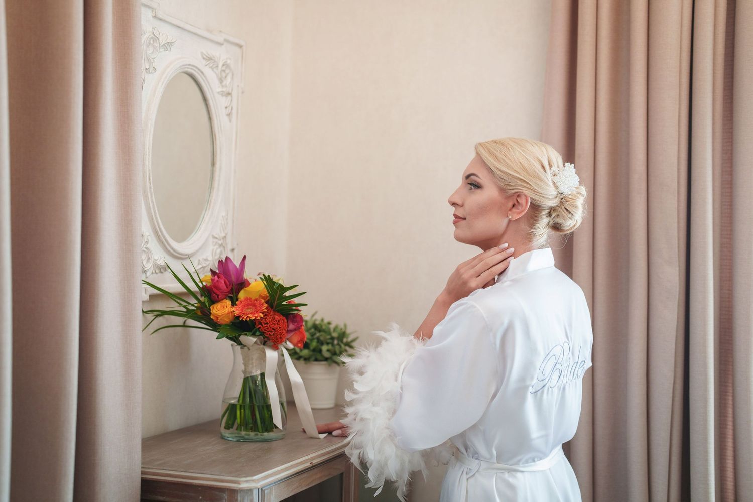 A bride in a white robe admires a colorful floral arrangement next to an ornate wall mirror.