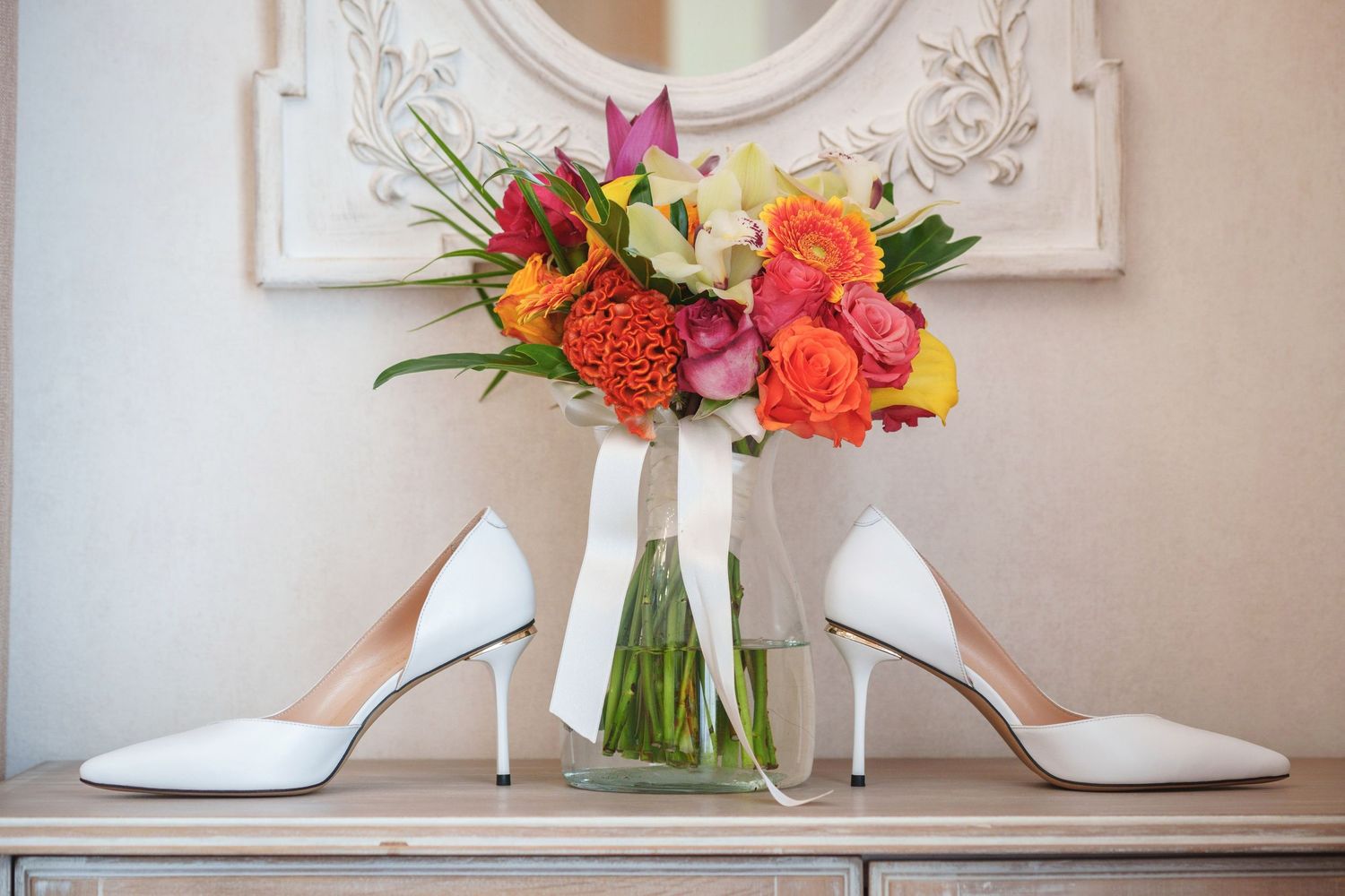 White bridal heels frame a vibrant bouquet of orange and pink flowers in a glass vase.