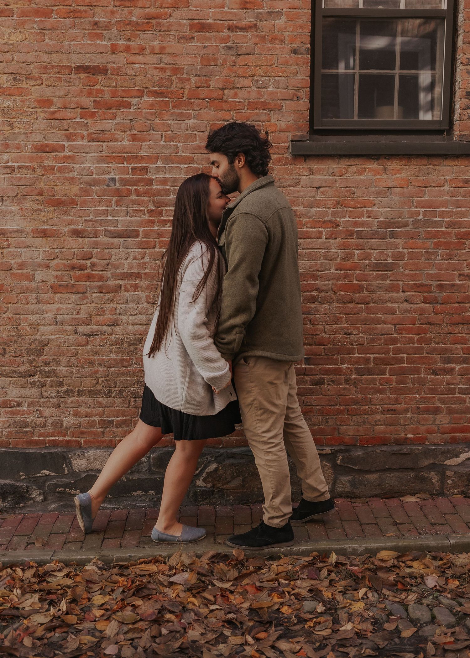 A couple shares an intimate moment against a rustic brick wall with autumn leaves scattered on the ground in Rhode Island.