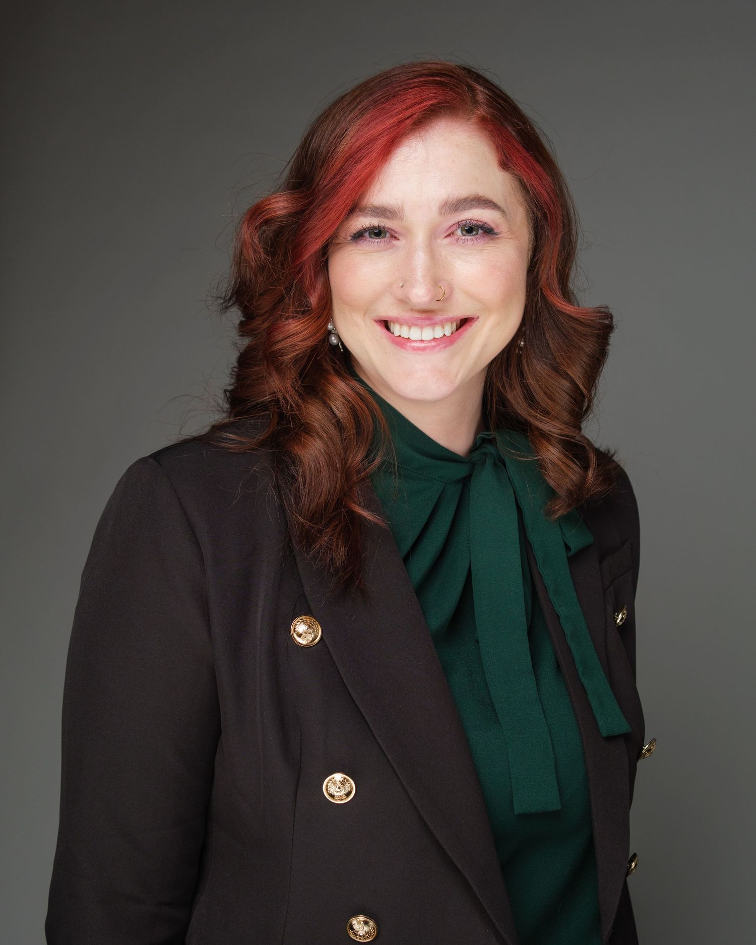 Professional headshot in a black blazer with gold buttons and green bow blouse against a dark gray background.
