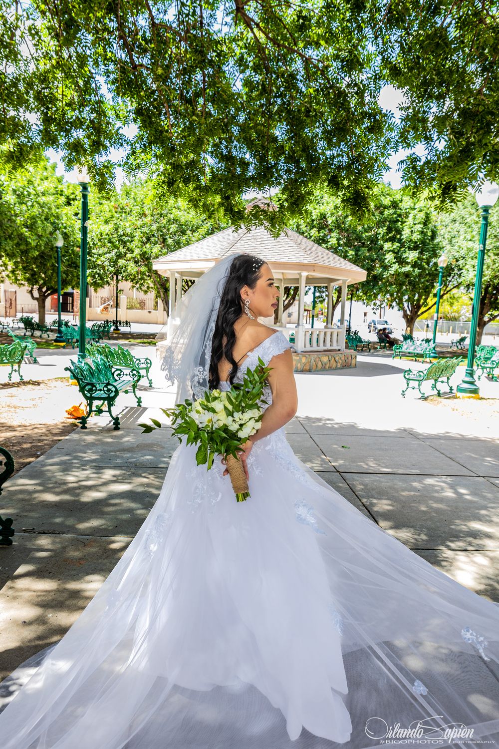 A bride spins her dress in a sunlit courtyard surrounded by green trees and park benches.