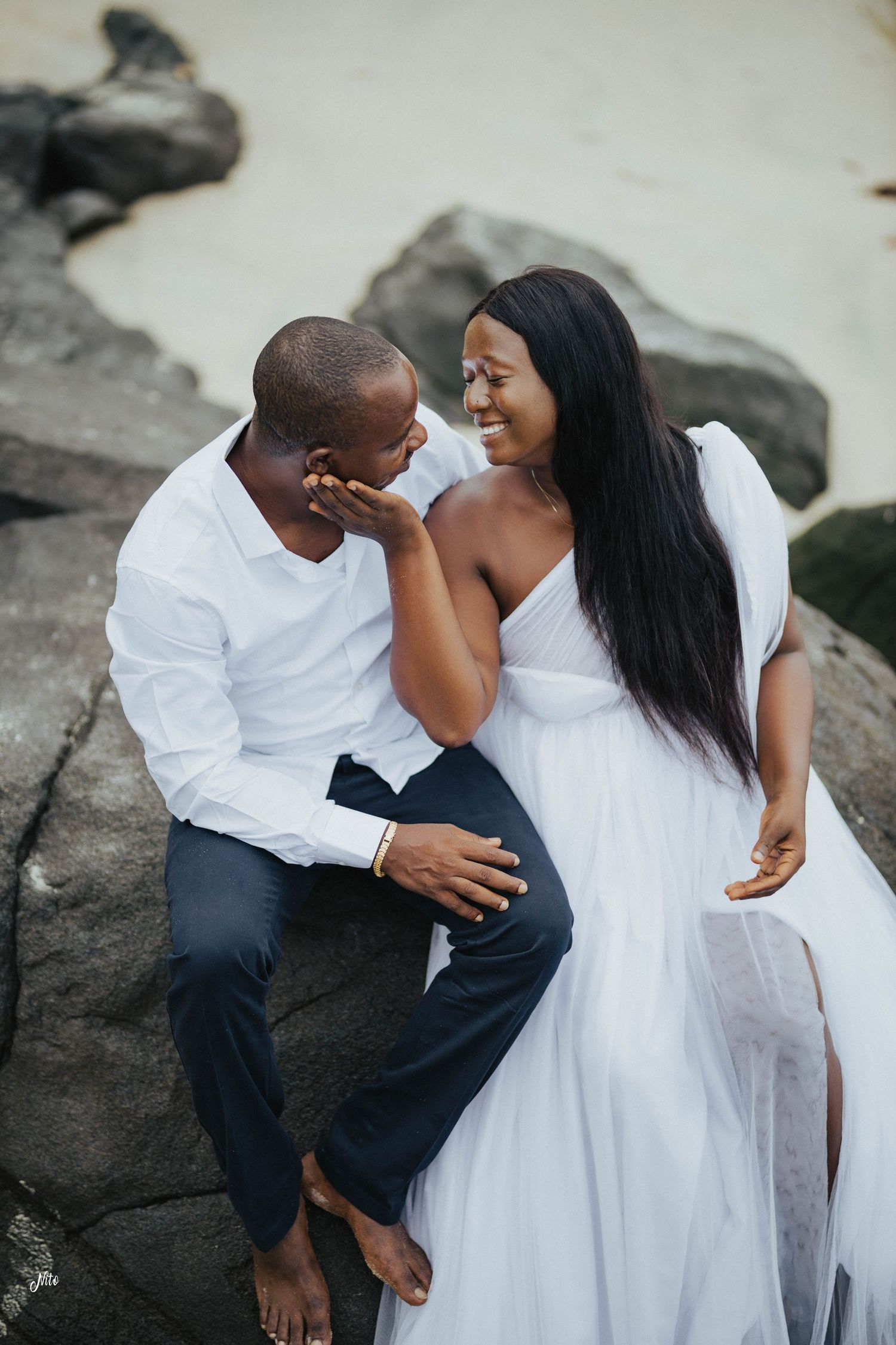 Intimate moment shared between couple in white attire sitting on coastal rocks.