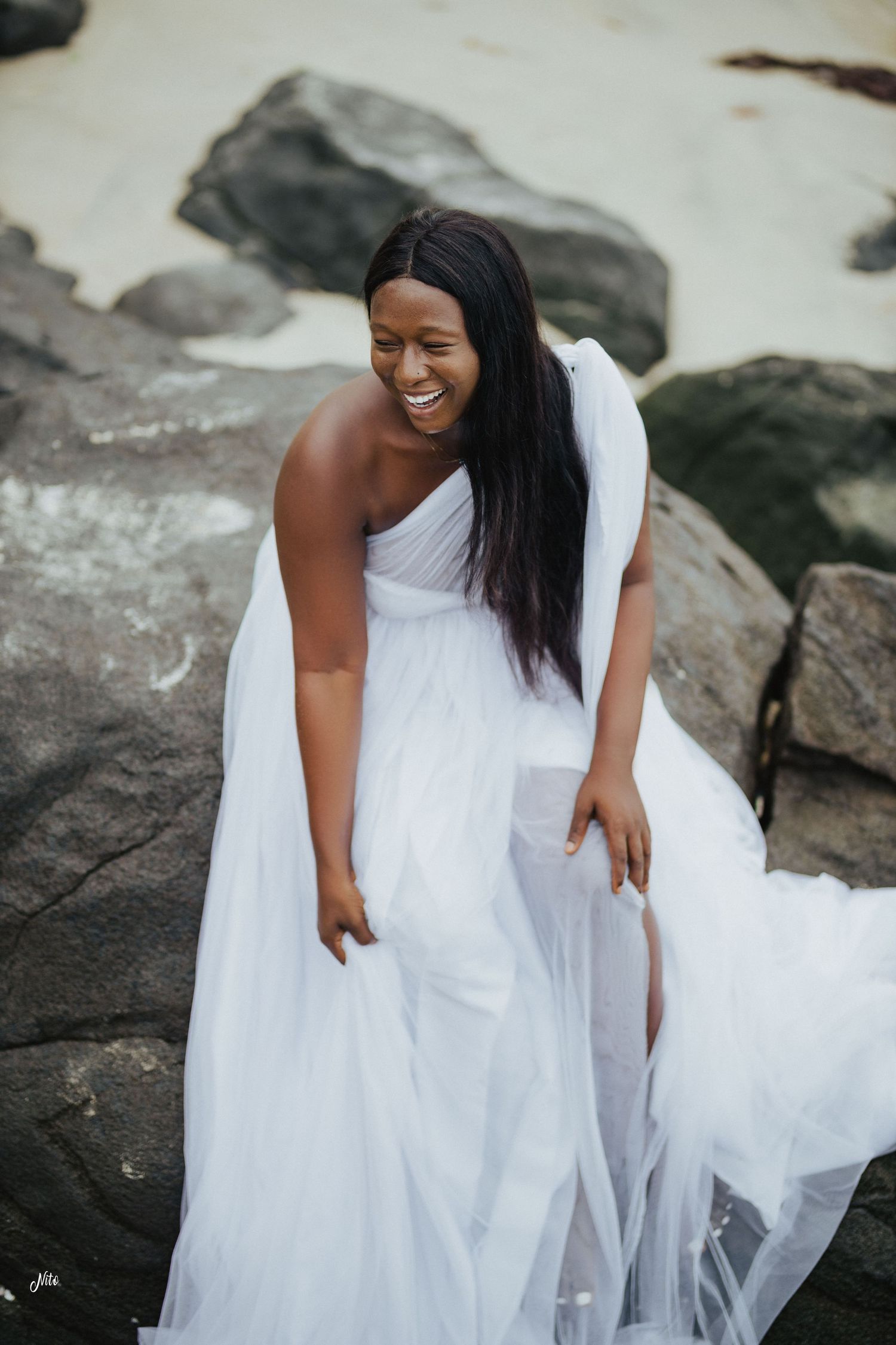 Beautiful bride in a flowing white dress poses on rocky coastal terrain.