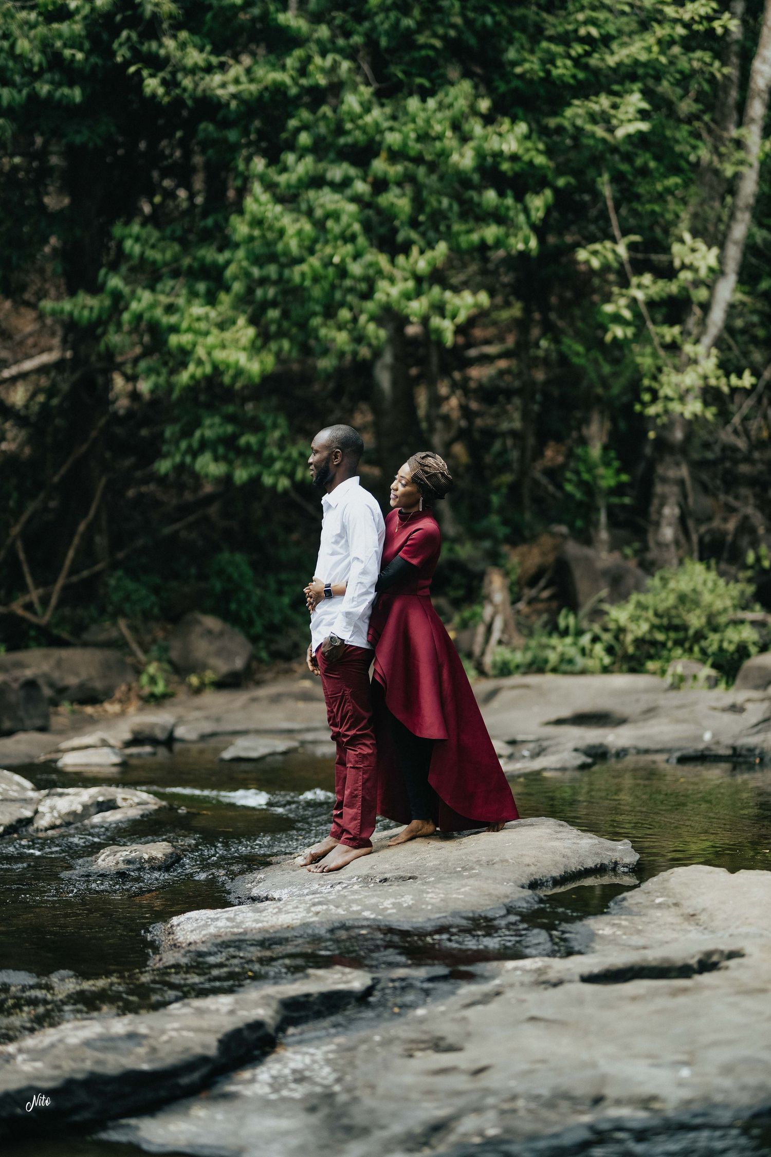 A couple standing back-to-back on rocks in a forest stream.