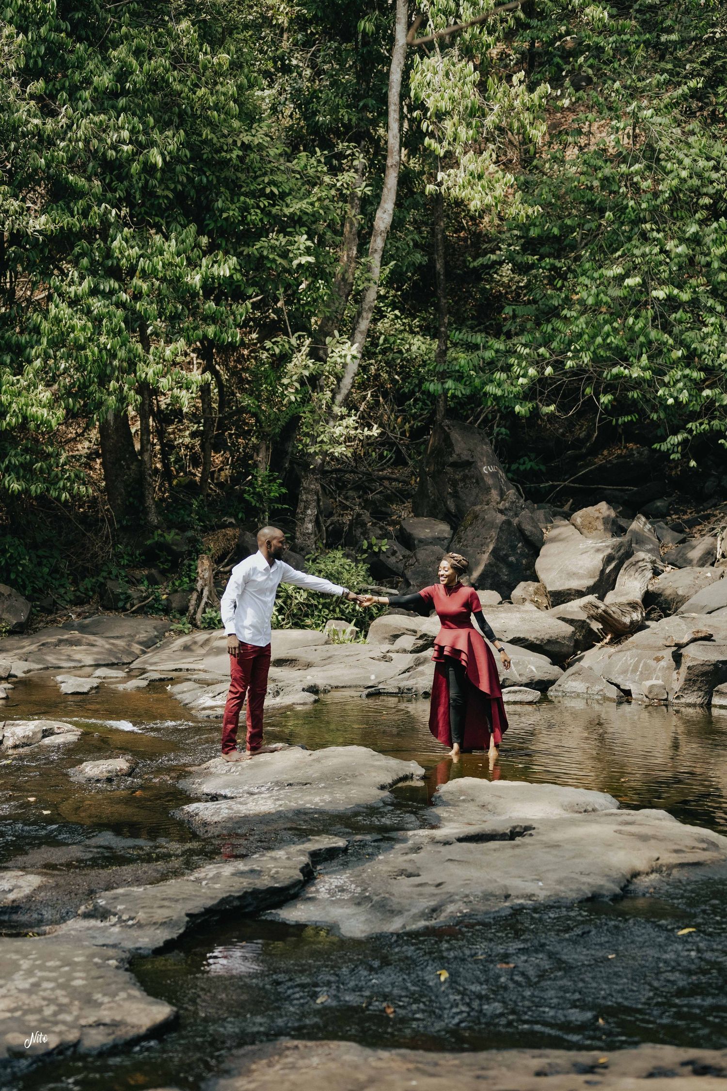 A couple balancing on rocks near a stream in a forest setting.