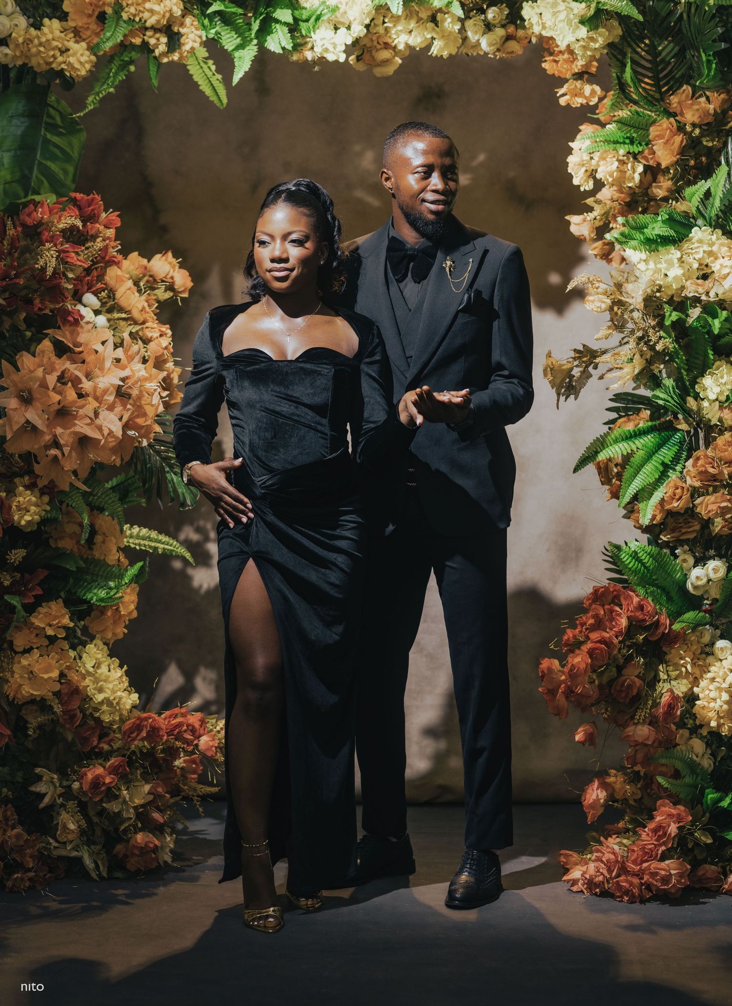 Elegant couple poses in formal black attire against floral backdrop with hydrangeas and greenery.