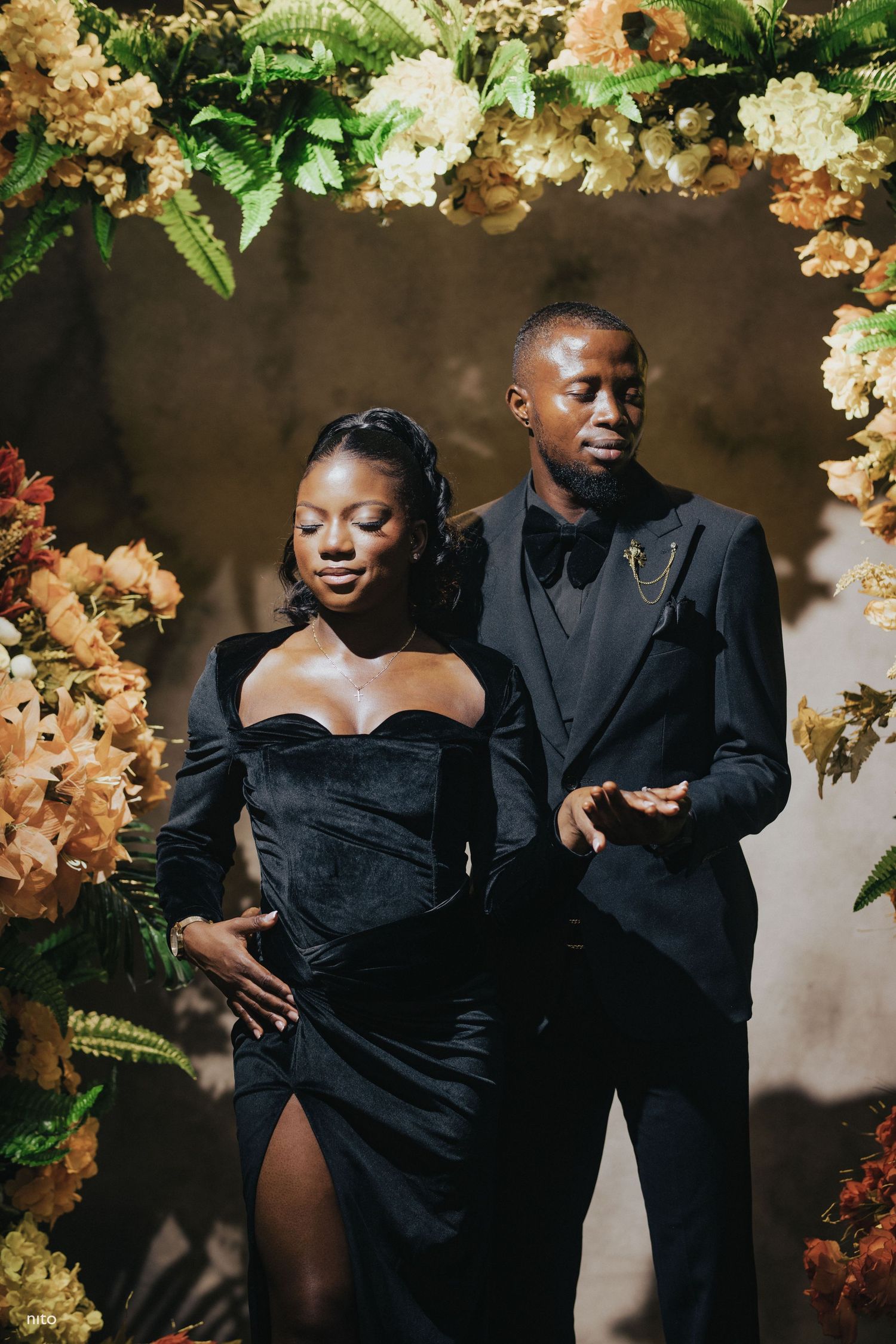 Stylish pair in black formal wear share playful moment amid floral arch decorations.