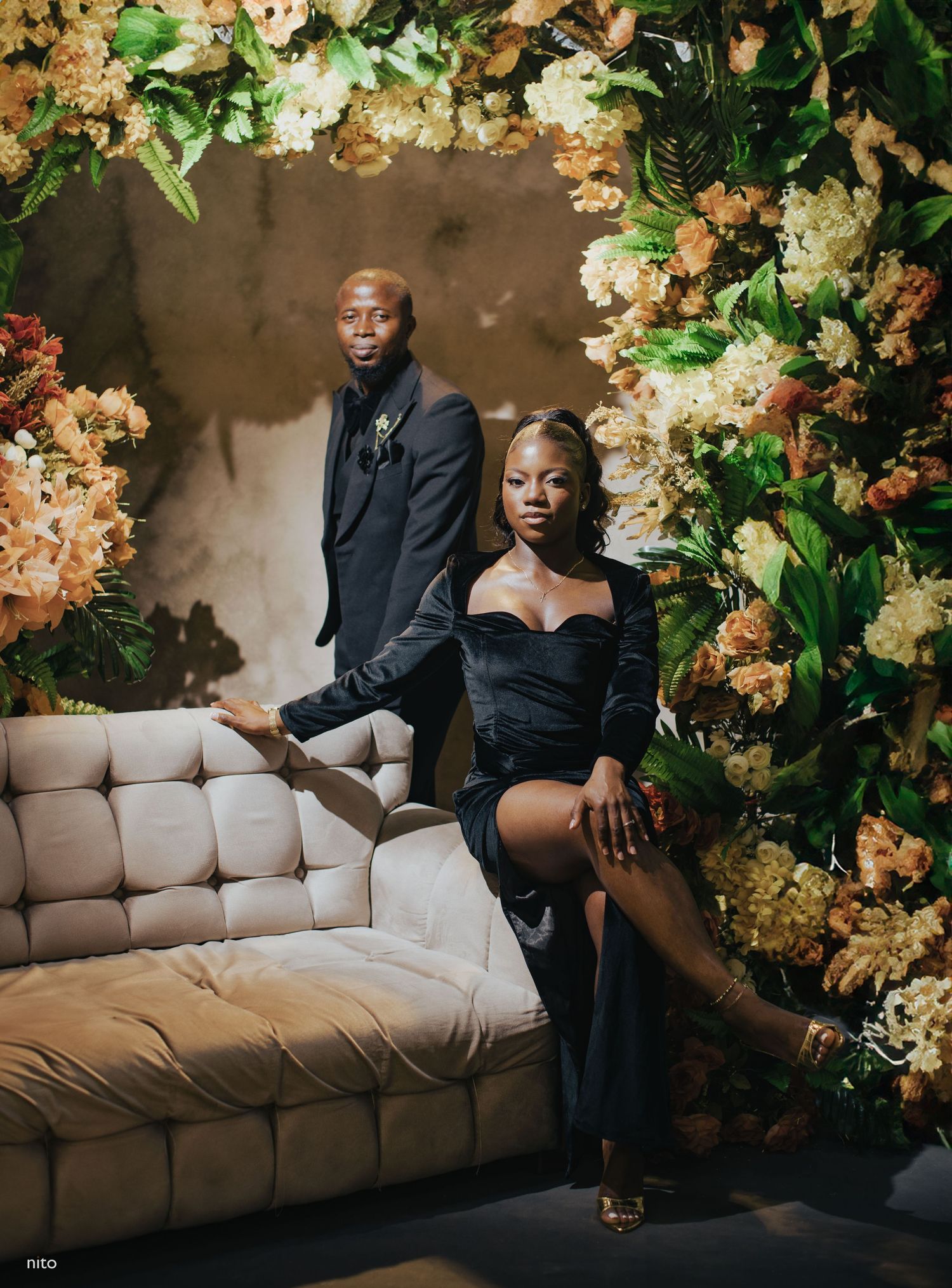 Fashionable duo poses on beige couch surrounded by luxurious floral arrangements.