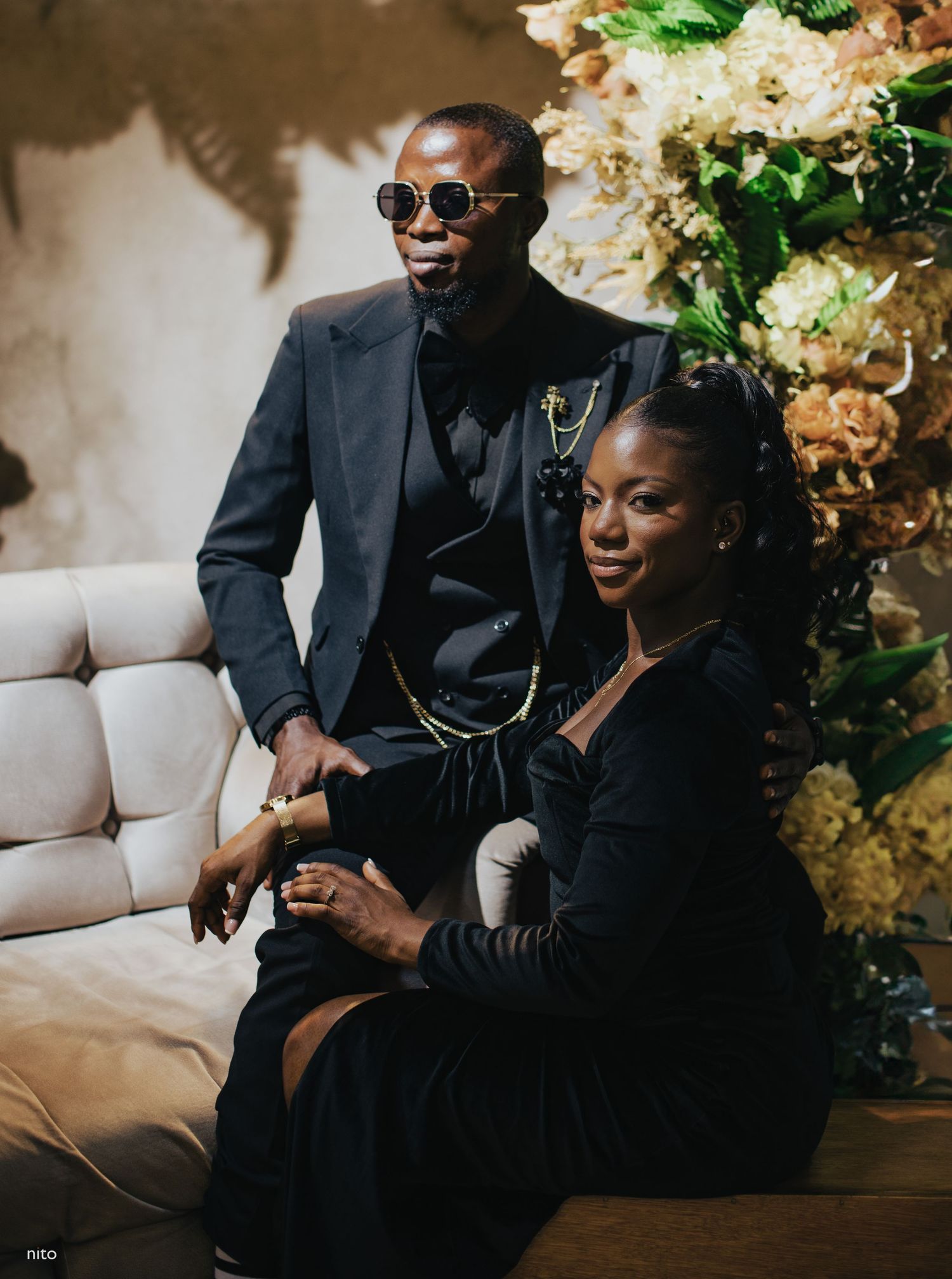 Couple in black formal wear relaxes on couch with sunglasses and floral decor.