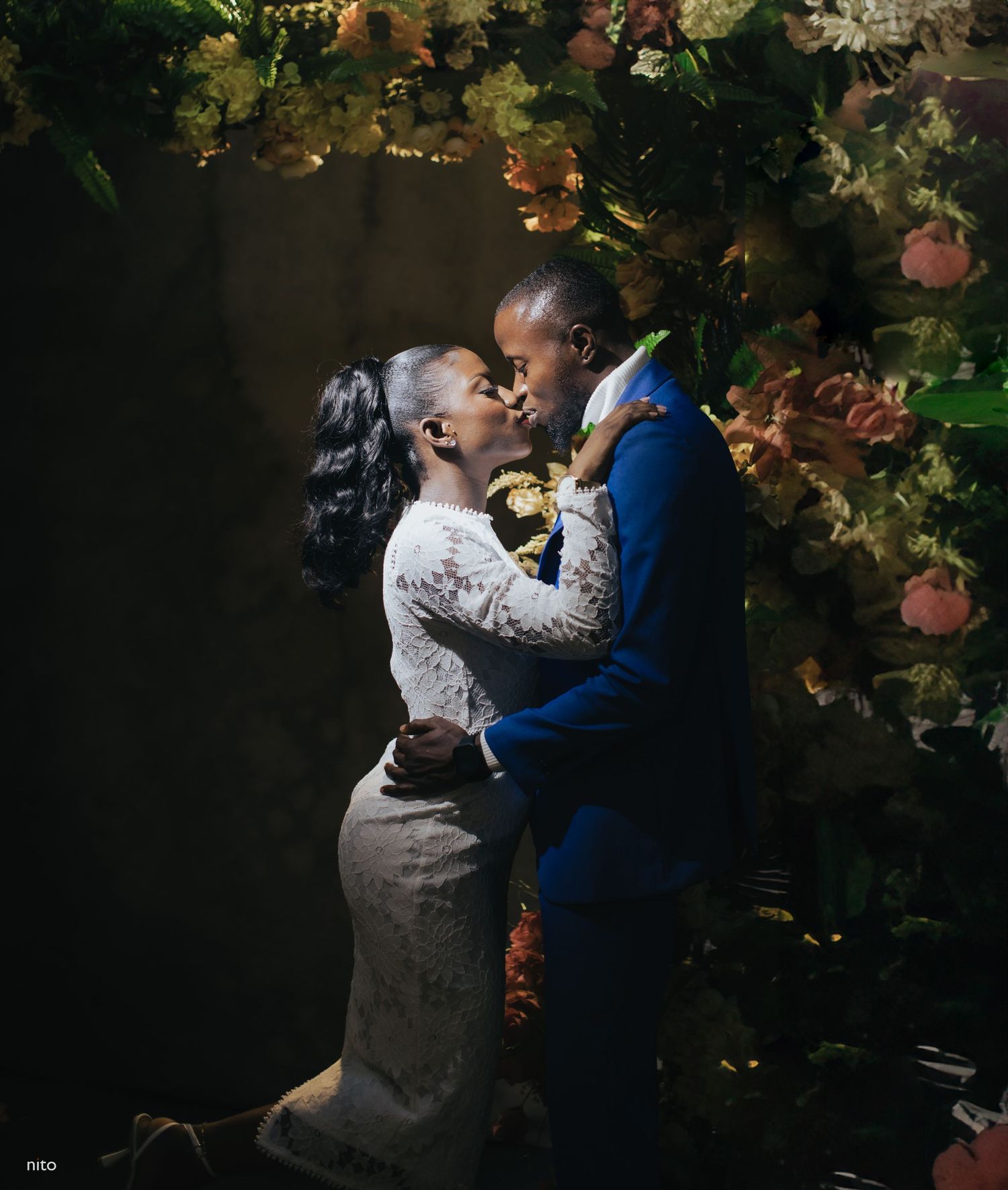 Romantic embrace between couple in formal wear under floral arch with dramatic lighting.