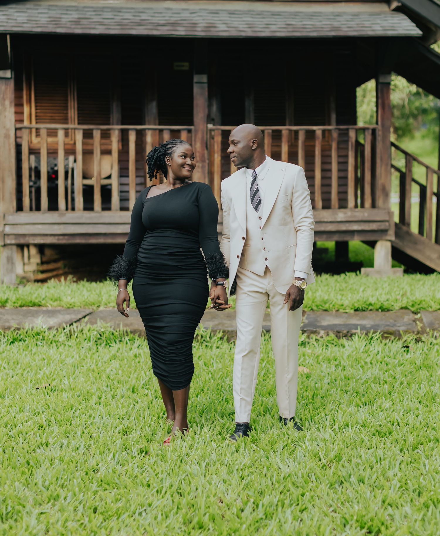 Stylish couple in formal wear holding hands while standing on grass in front of a rustic cabin.