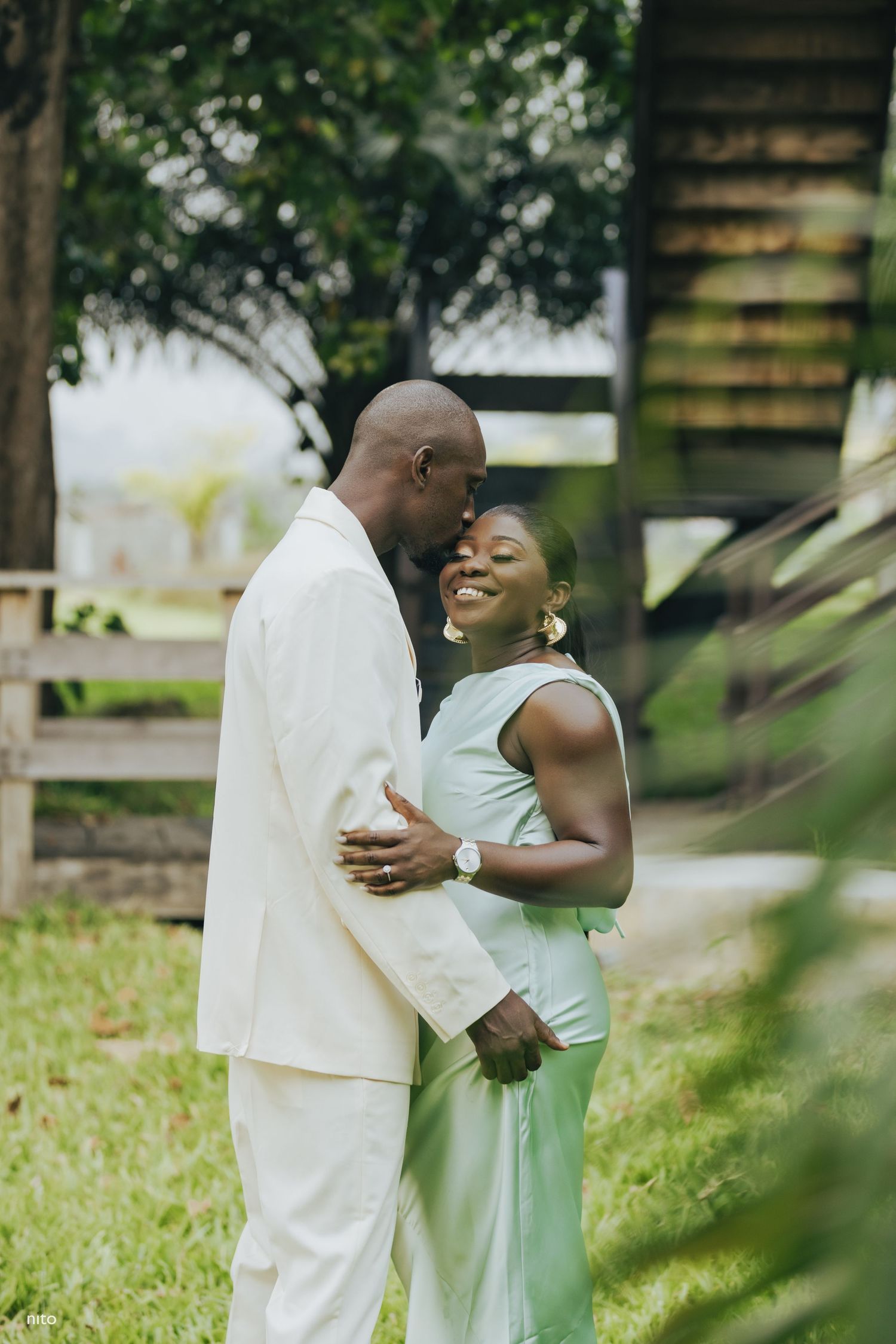A tender moment between a couple in white and mint green outfits against a rustic backdrop.