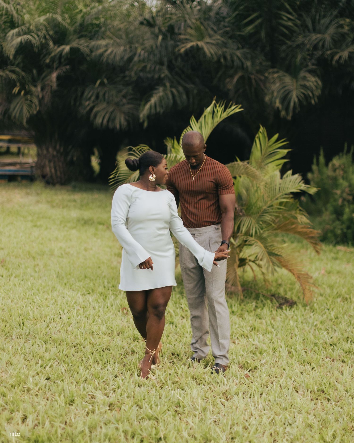 A loving couple shares an intimate moment while standing on grass with tropical foliage behind them.