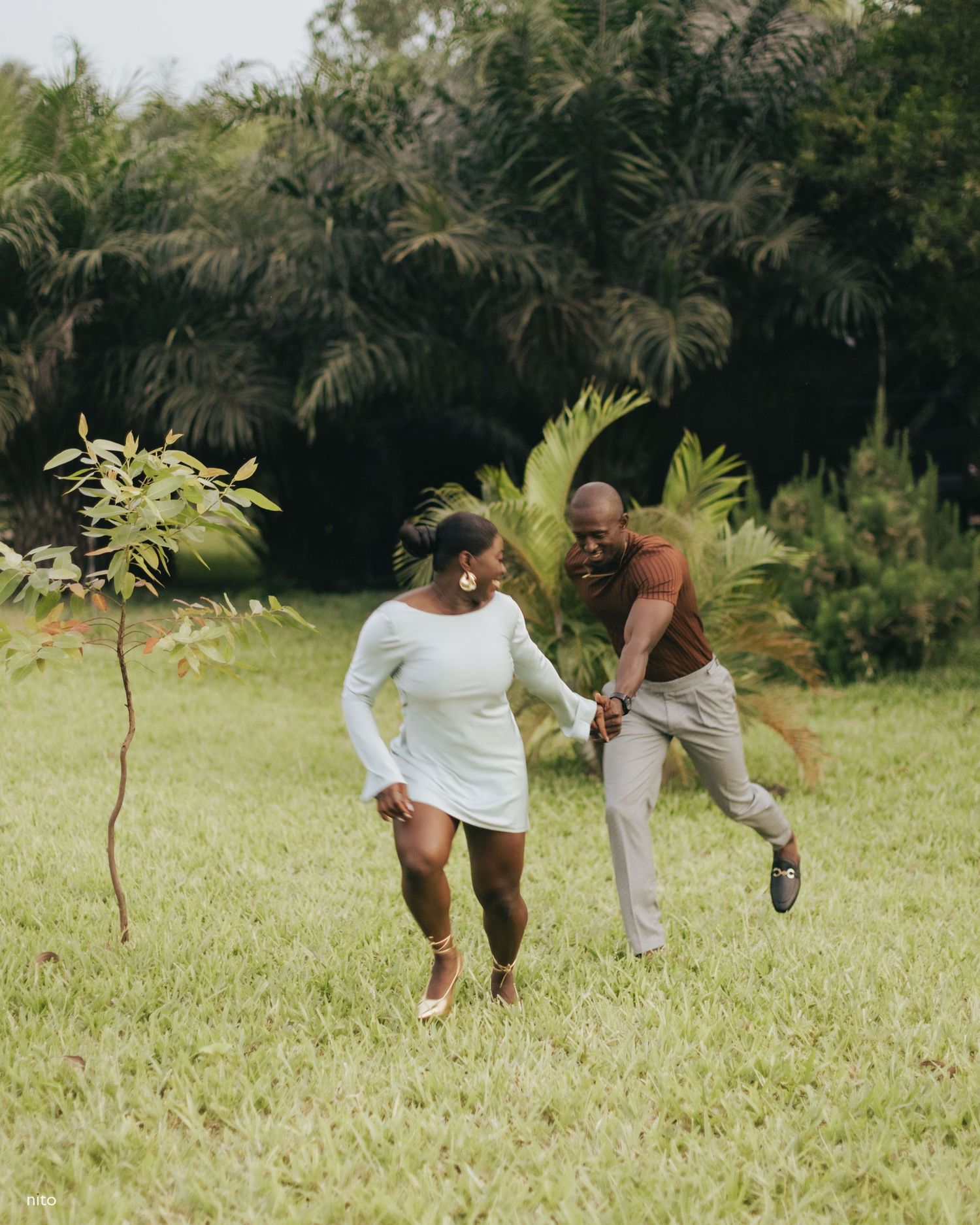 Two people running and laughing together on grass with tropical plants in the background.