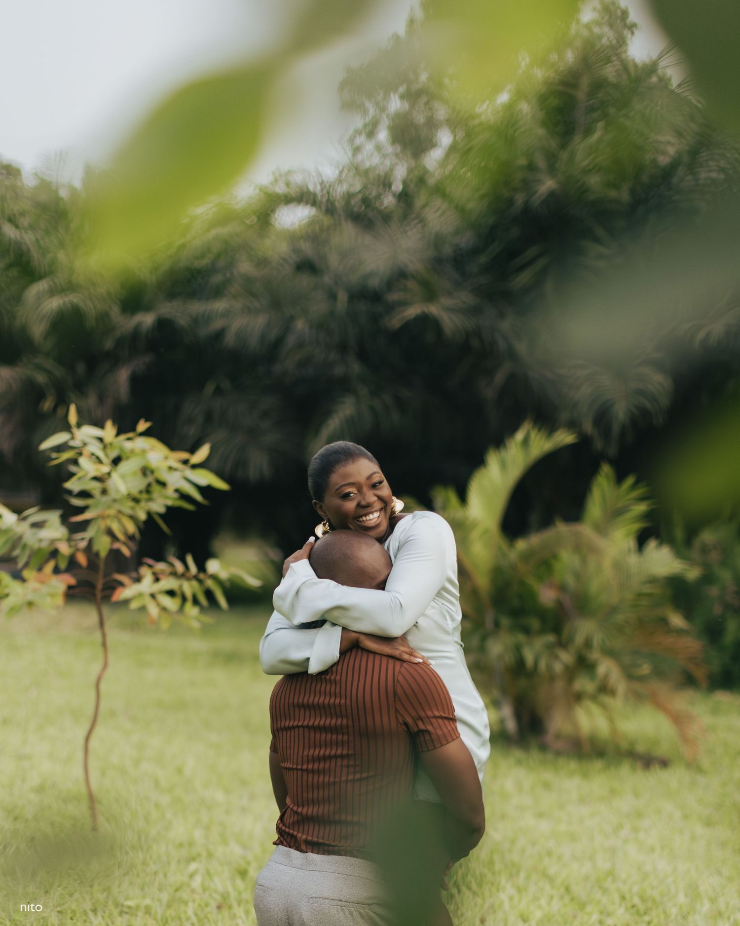A couple embraces joyfully in a garden setting with lush greenery and palm trees.
